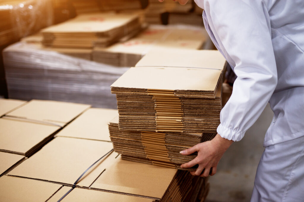 corrugated packaging suppliers — Close up of young female worker picking up stacks of folded cardboard boxes from a bigger stack in factory storage room.